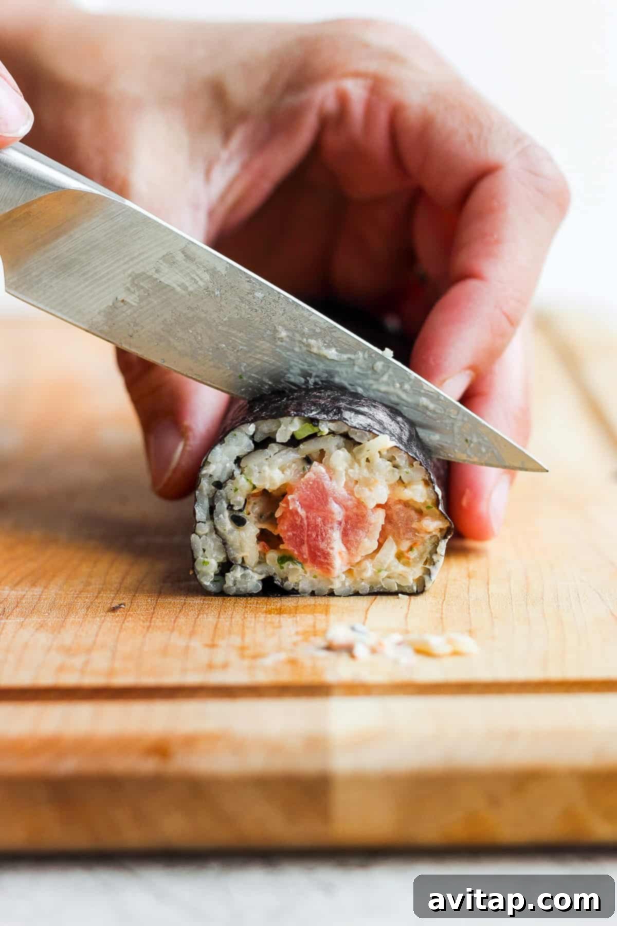 A sharp knife neatly slicing through a freshly prepared spicy tuna roll on a wooden cutting board.
