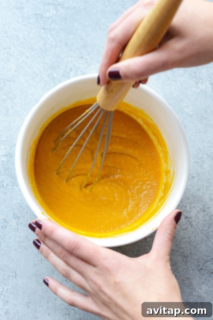 Rich orange pumpkin puree being vigorously whisked in a clear glass bowl, showcasing its smooth, creamy texture.