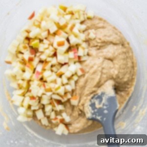 Diced apples being carefully folded into the muffin batter with a spatula in a large mixing bowl.