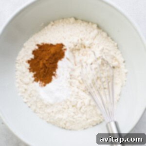 A bowl containing dry muffin ingredients, including flour, cinnamon, and a whisk, ready for mixing.