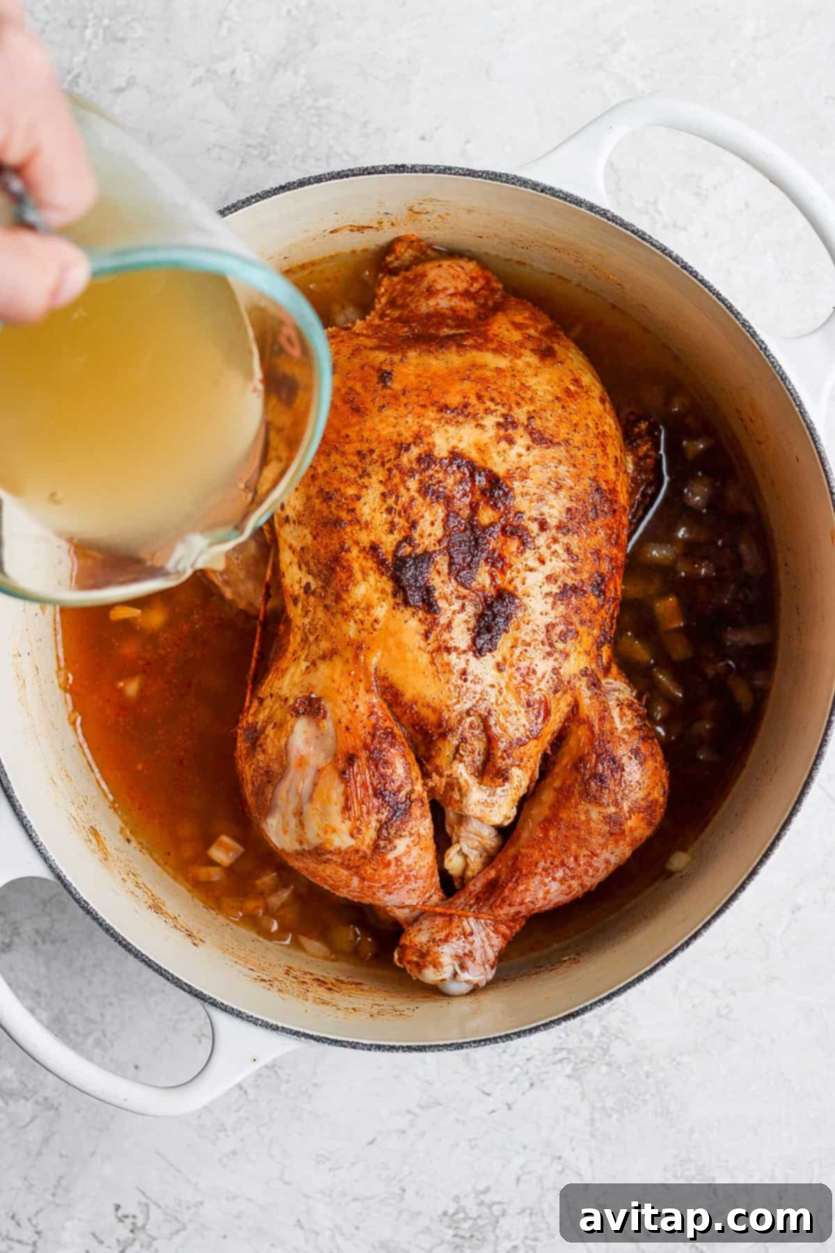A person pouring chicken broth over a whole roasted chicken in a Dutch oven, preparing it for the oven.