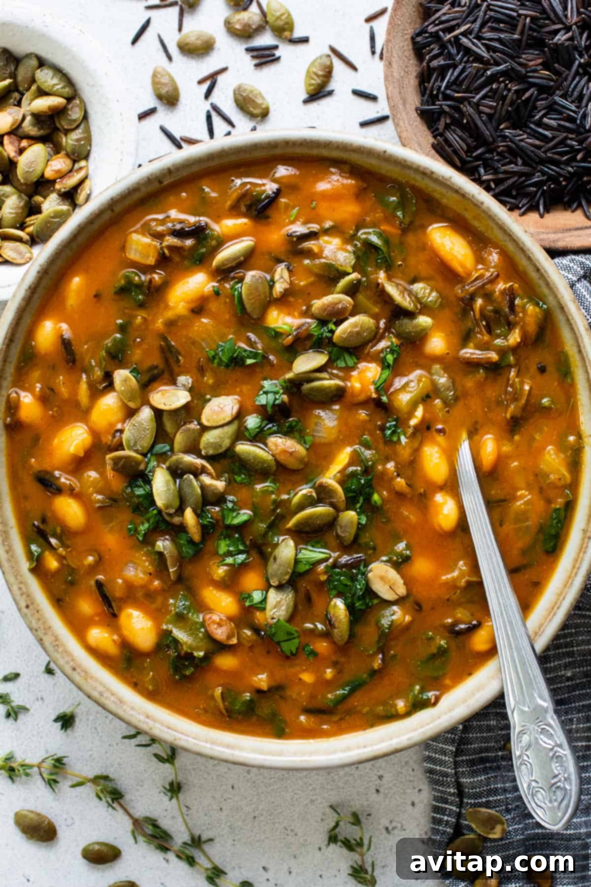 A steaming bowl of creamy pumpkin wild rice soup, garnished with pumpkin seeds and white beans, on a rustic surface.