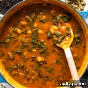 A pot of hearty soup with wilted kale and a wooden spoon inside.