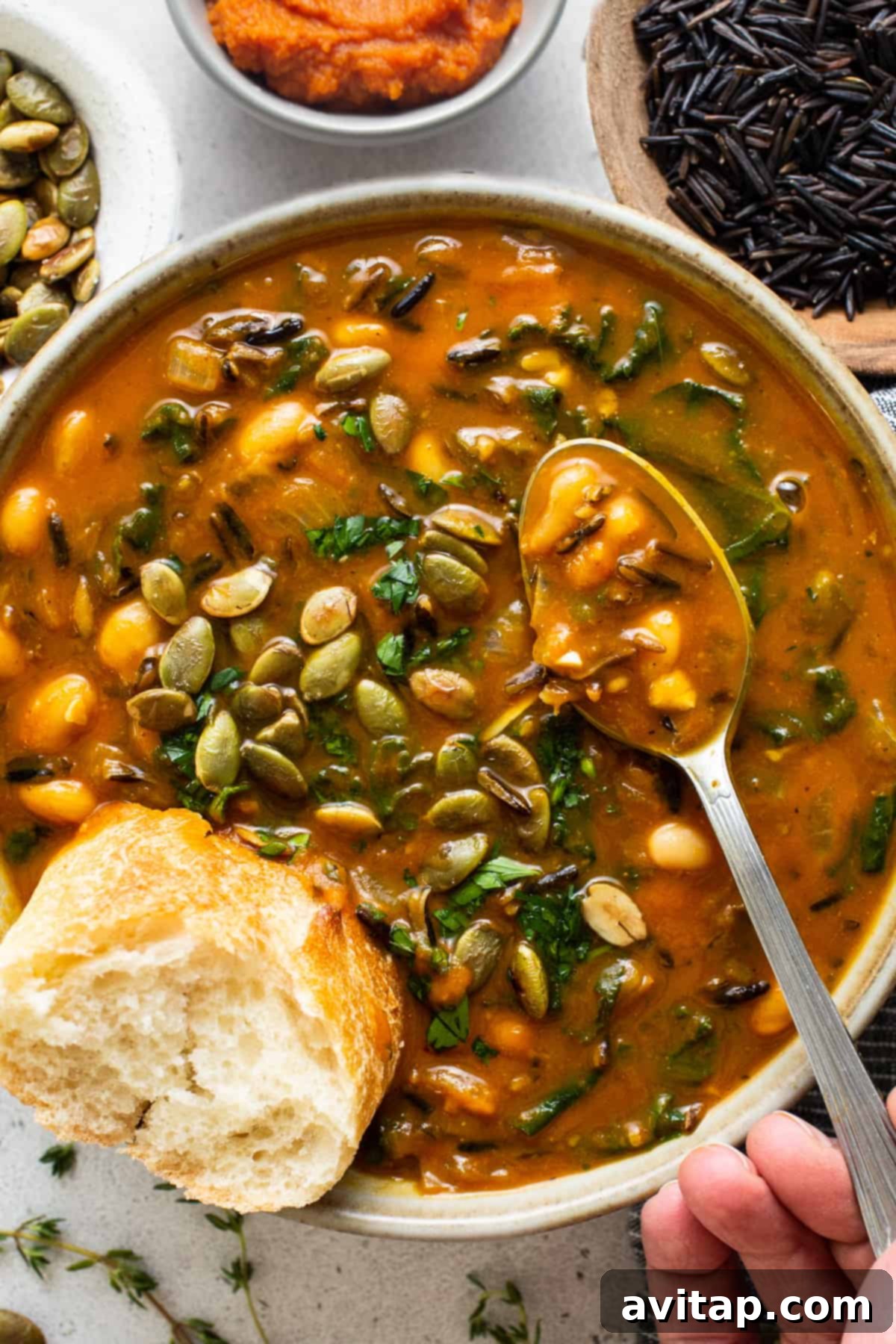 A hand holding a spoon, scooping creamy pumpkin wild rice soup from a bowl garnished with pepitas.