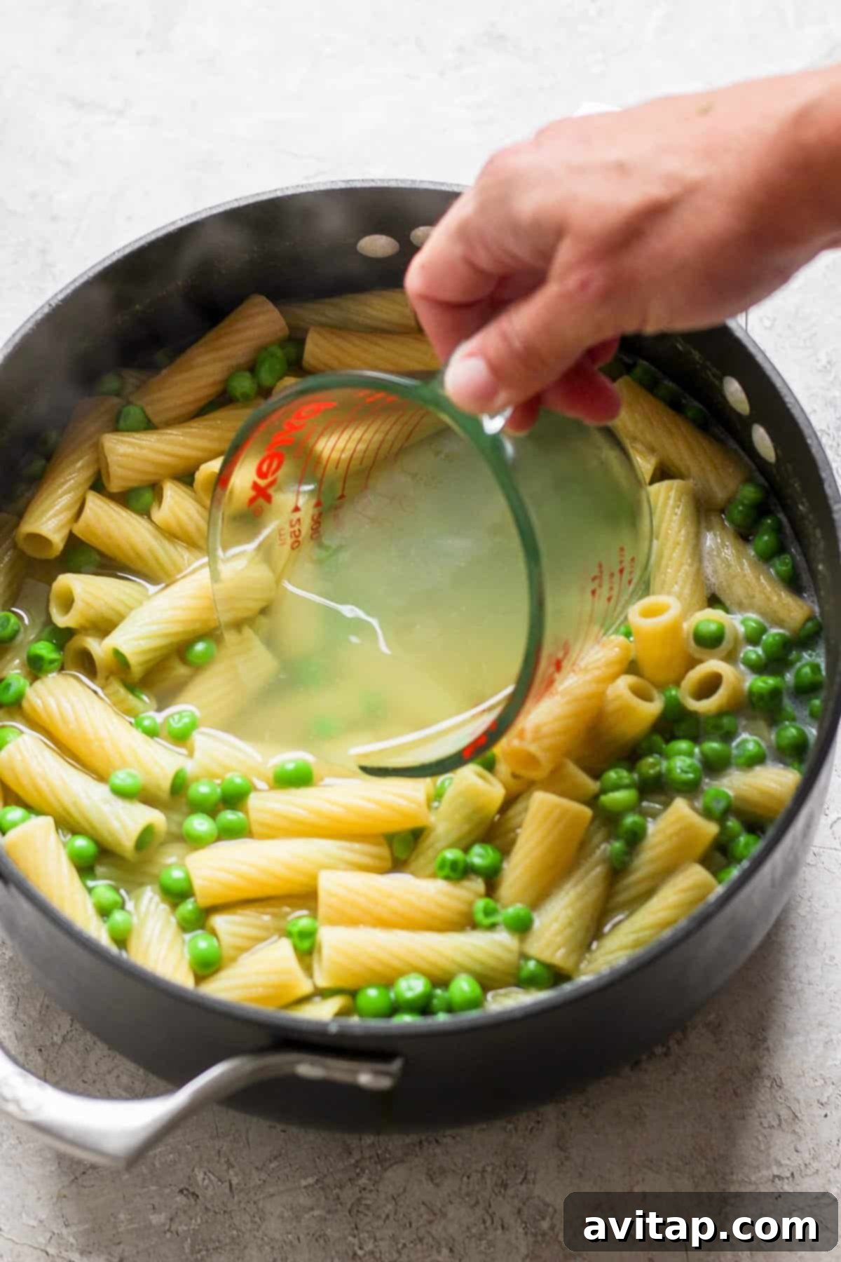 Creamy One-Pot Boursin Pasta 5 A person pouring pasta and peas into a pan, illustrating the one-pot cooking process.
