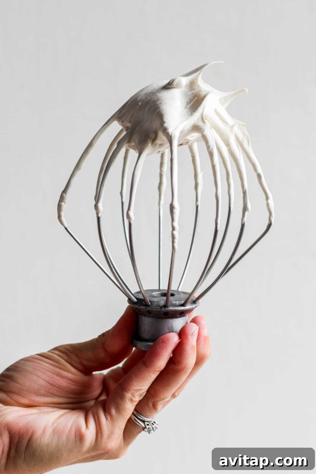 A person holding a whisk coated in fluffy, light brown meringue, ready to top a dessert.