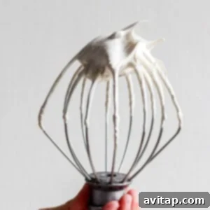 A person holding a whisk with a generous amount of brown sugar Swiss meringue on it, demonstrating its thick and fluffy texture.