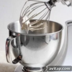 A stand mixer vigorously whisking the meringue mixture, showing it transforming into fluffy, stiff peaks.