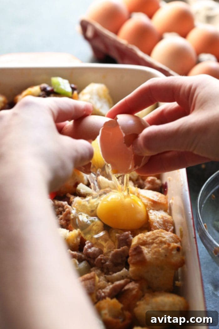 A close-up view of hands gently cracking fresh, large eggs directly over a prepared spicy Italian breakfast casserole mixture, which includes colorful vegetables, sausage, and bread, nestled in a baking dish, symbolizing the final preparation stage before baking.