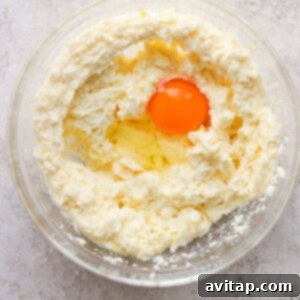 A single egg is visible in a bowl of creamy cookie dough mixture, being gently beaten in until fully combined.