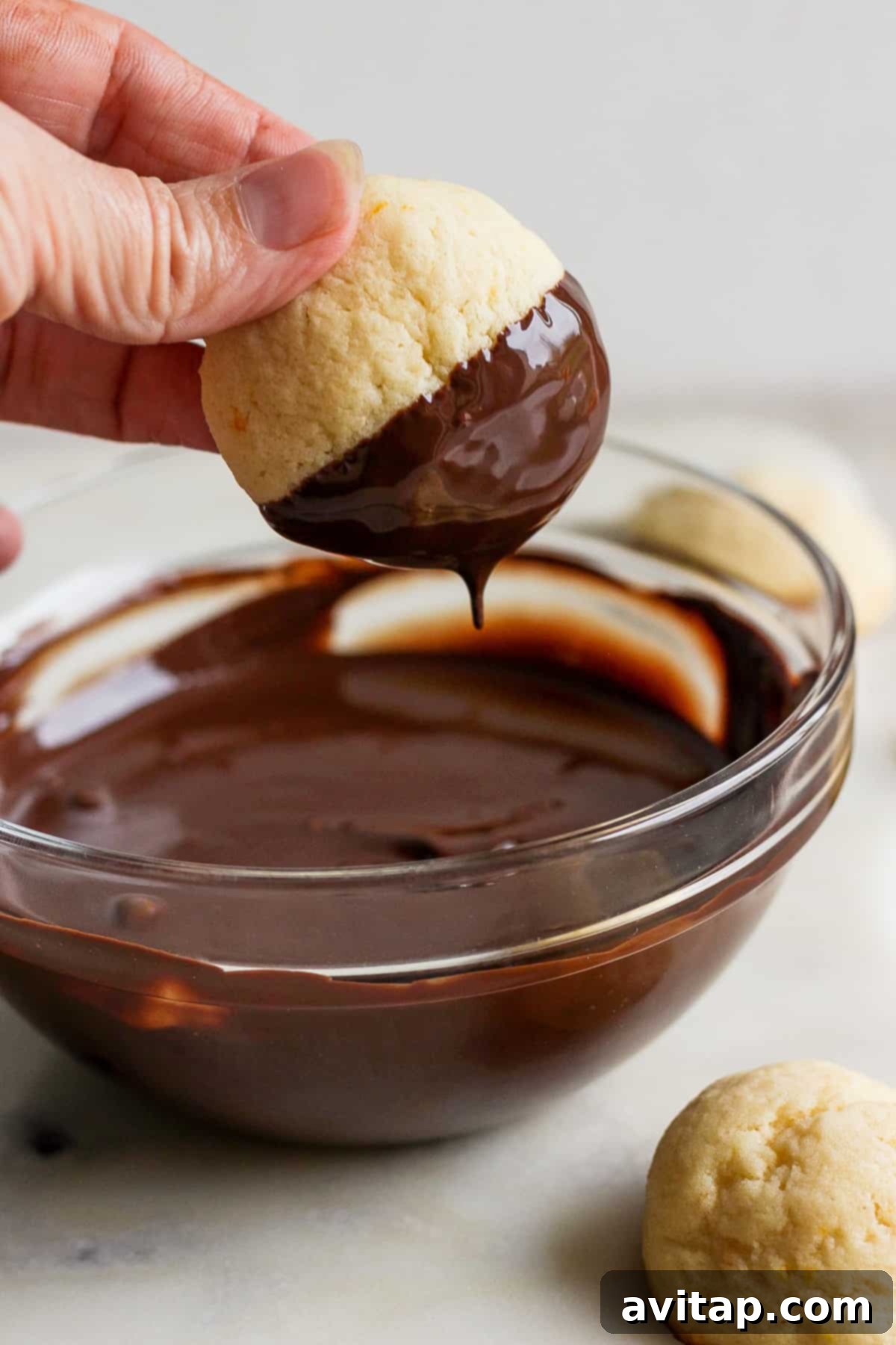 Orange Kissed Chocolate Ricotta Clouds 5 A hand gently dips a freshly baked ricotta cookie into a bowl of glossy, melted dark chocolate, preparing it for its final touch.