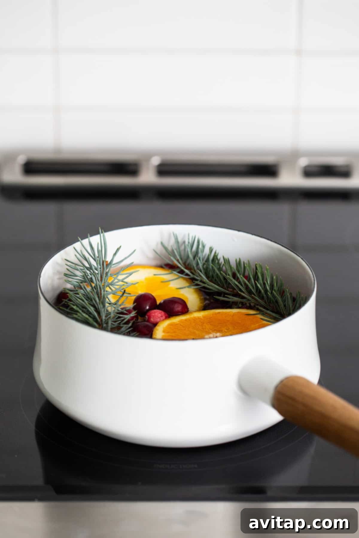 A crisp white pan filled with bright orange slices and fresh green pine needles, gently simmering on a stovetop, creating a warm, inviting atmosphere.