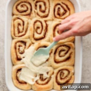 A person is dipping a spatula into a dish of cinnamon rolls.