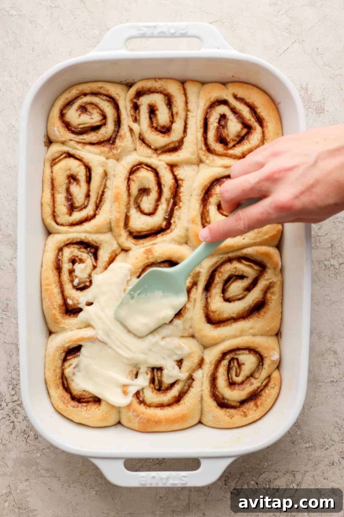 A person is dipping a spatula into a dish of cinnamon rolls.