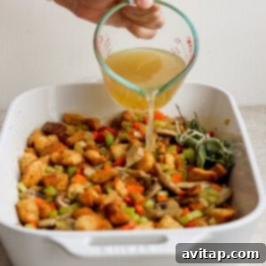 A person pouring broth over a dish of stuffing and vegetables in a casserole dish.
