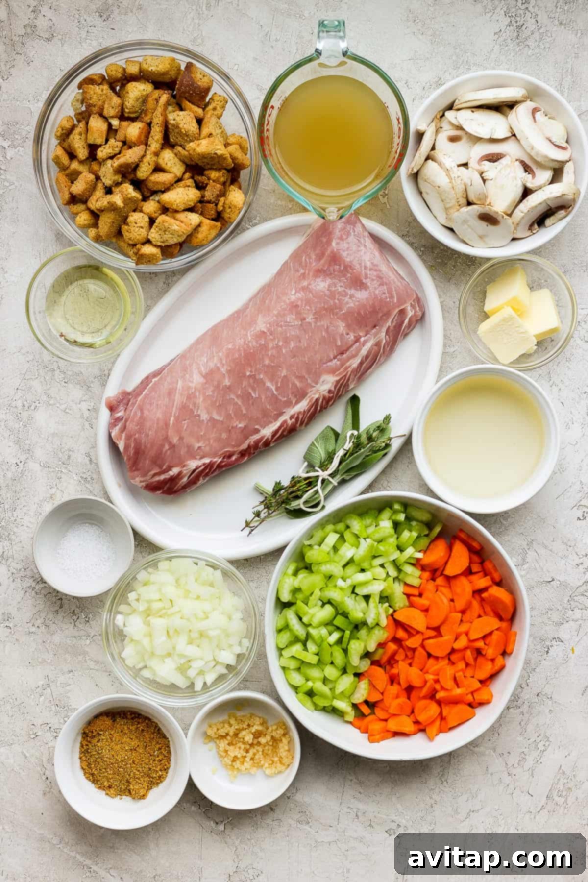 An array of fresh ingredients for roasted pork tenderloin and stuffing, including tenderloin, carrots, mushrooms, and herbs, arranged in white bowls on a pristine white background.