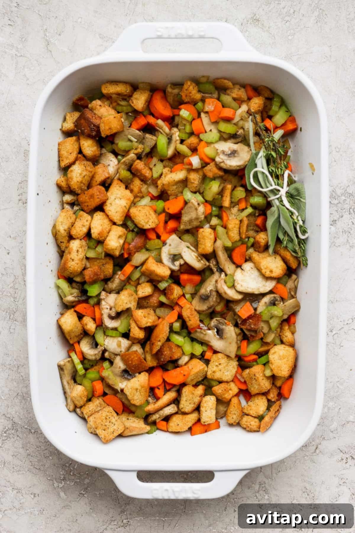 A white baking dish brimming with a flavorful stuffing mixture of stale bread, sautéed vegetables, and aromatic herbs, ready for the pork tenderloin.