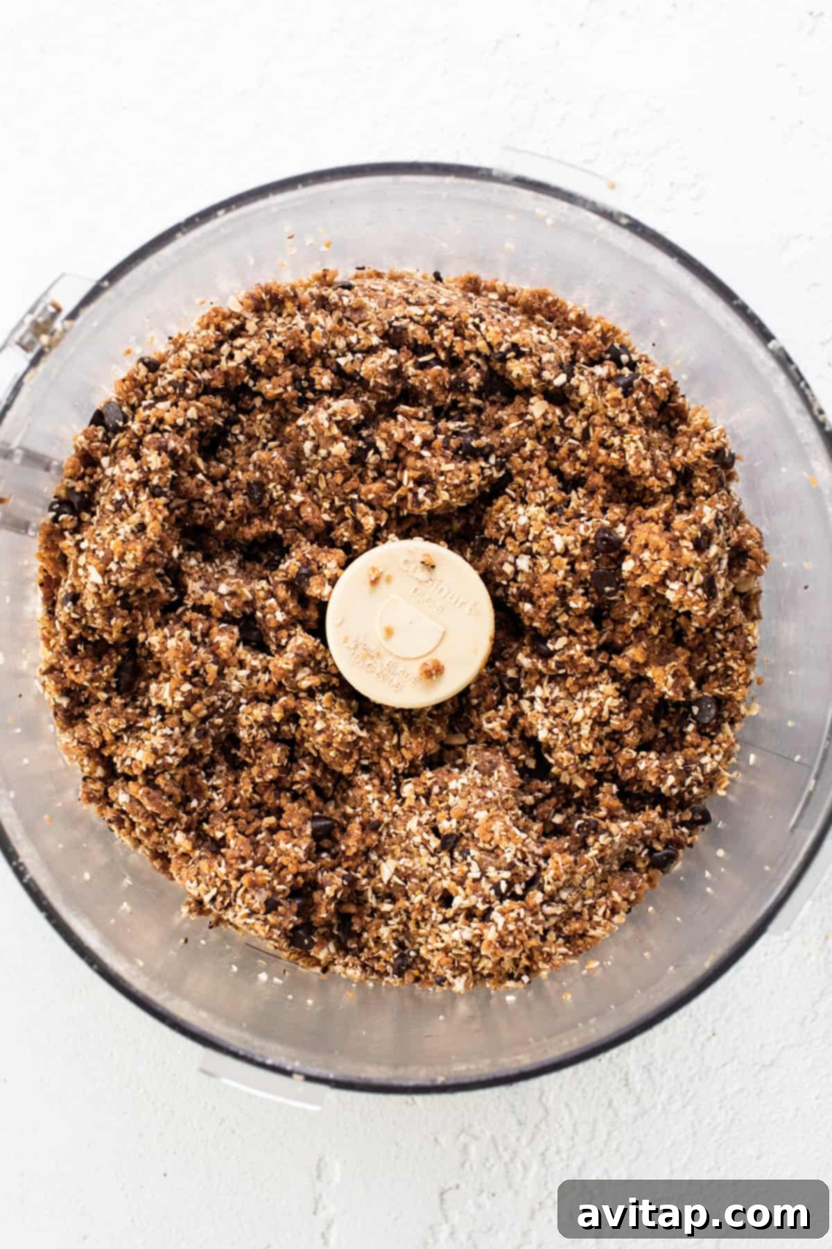 A food processor containing a blend of granola-like ingredients and a banana, illustrating the mixing process for the cookie dough base.