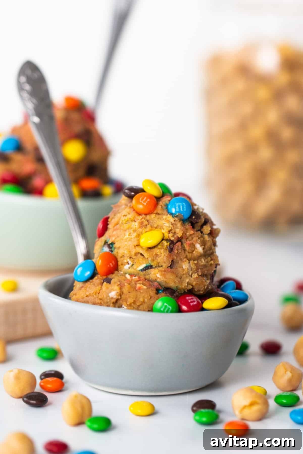 Protein Packed Monster Cookie Dough 7 A pristine white bowl overflowing with delicious protein monster cookie dough, generously topped with an array of colorful M&M's, with a spoon invitingly placed inside.