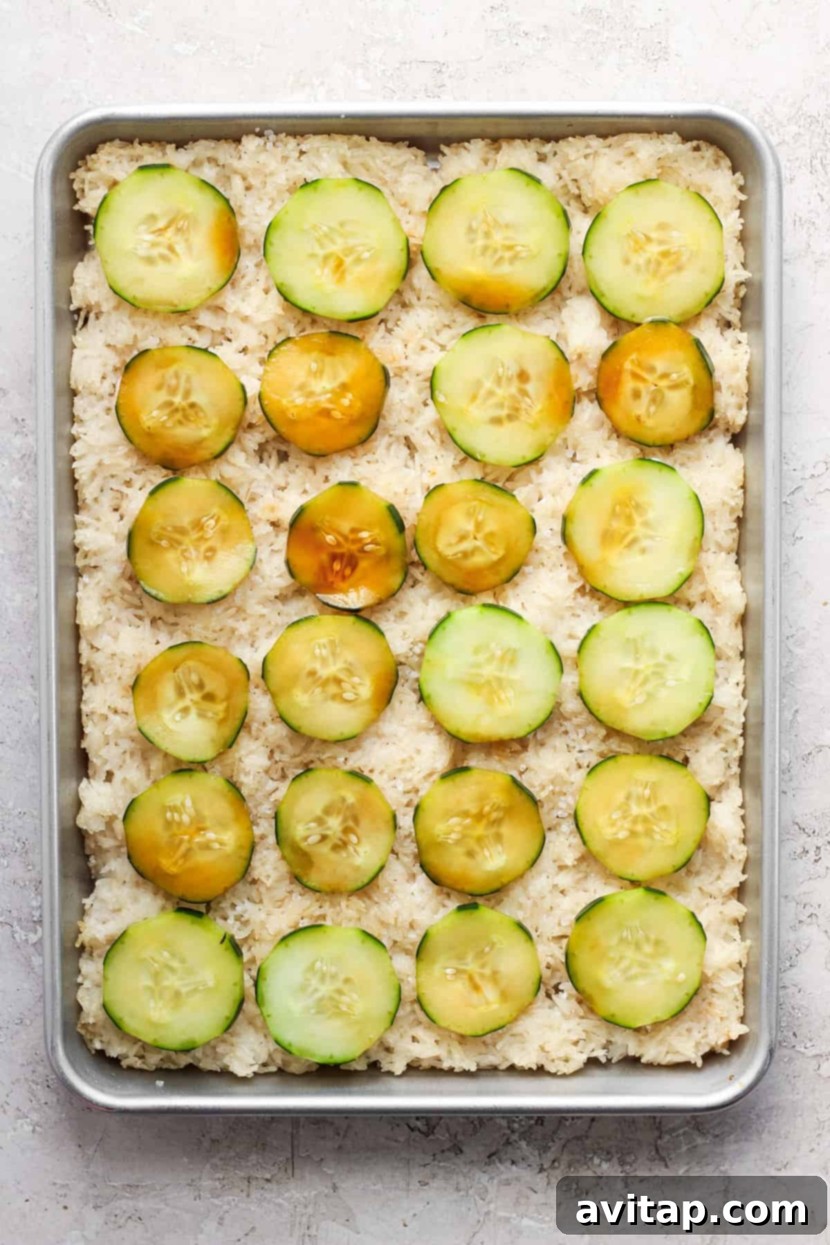 A sheet pan with crispy rice base topped with fresh cucumbers and ginger, ready for salmon.