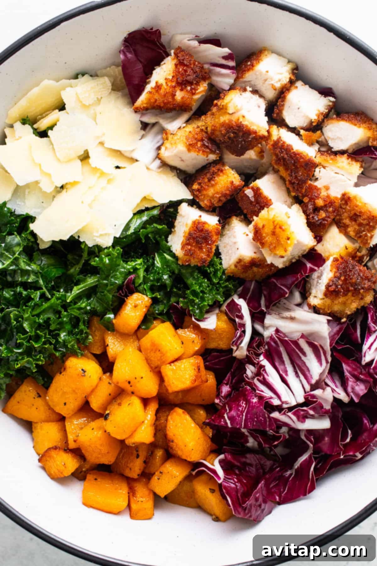 A bowl filled with fresh kale, diced crispy chicken, and shaved Parmesan cheese, ready for dressing.