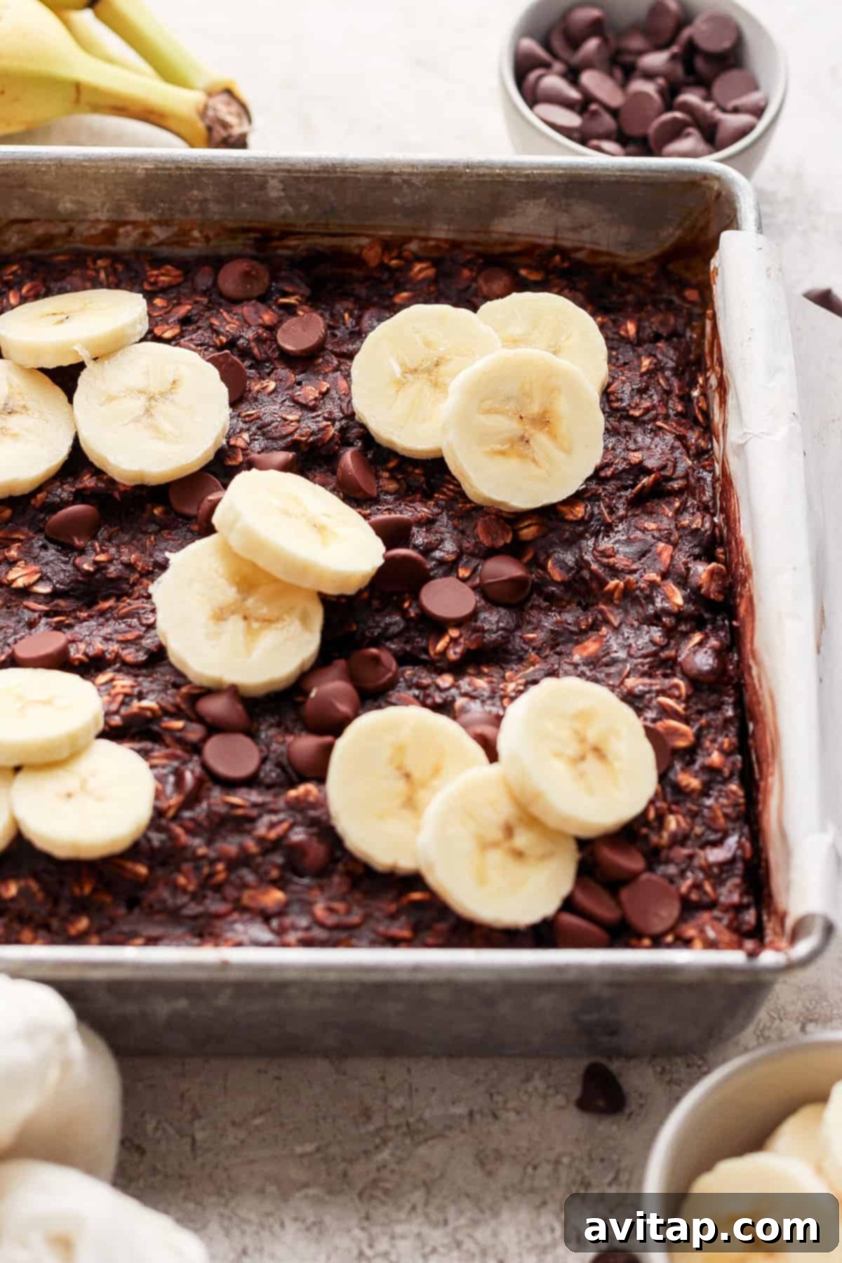 A baking pan filled with unbaked chocolate banana oatmeal, topped with extra banana slices and chocolate chips, ready for the oven.