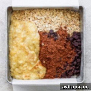 A deep baking dish filled with unbaked ingredients for chocolate banana oatmeal, featuring rolled oats, mashed bananas, and chocolate chips waiting to be mixed.