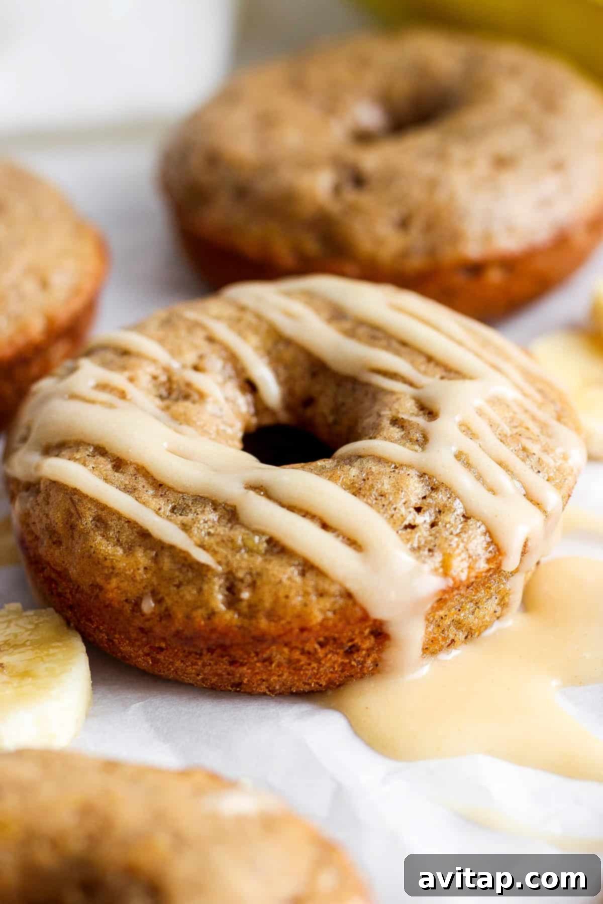 Banana Bread Ring Treats 2 A close-up image of a glazed banana doughnut, with more doughnuts and banana slices in the background.