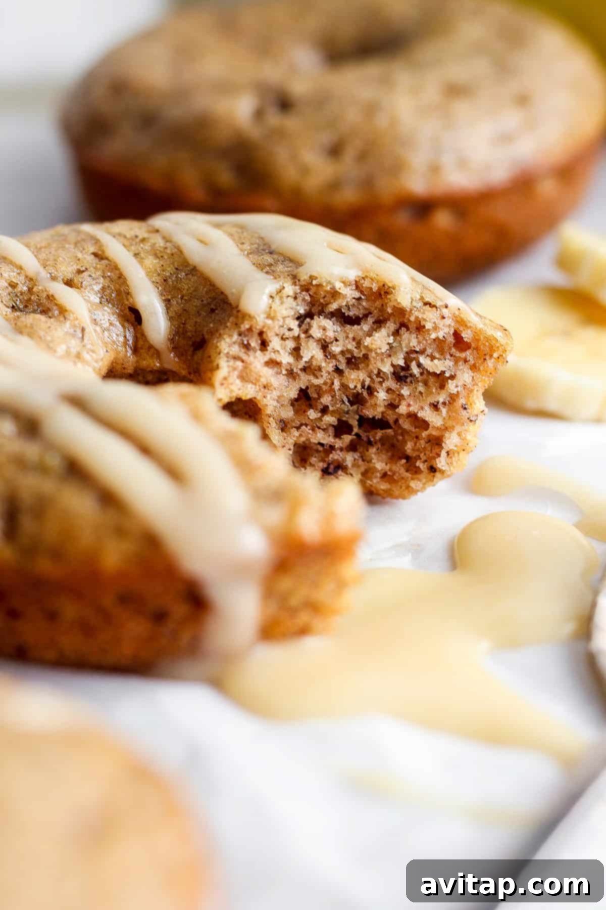 Banana Bread Ring Treats 7 Close-up of a partially eaten muffin with white glaze on top, with more muffins and banana slices blurred in the background.