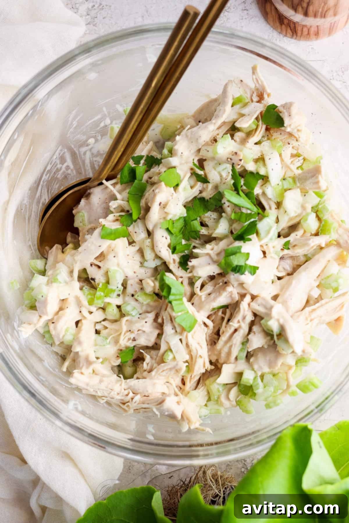 A bowl containing shredded chicken mixed with chopped celery and herbs, with a pair of gold-colored utensils.