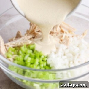 A creamy sauce is being poured into a bowl containing shredded chicken, diced onions, and chopped celery.