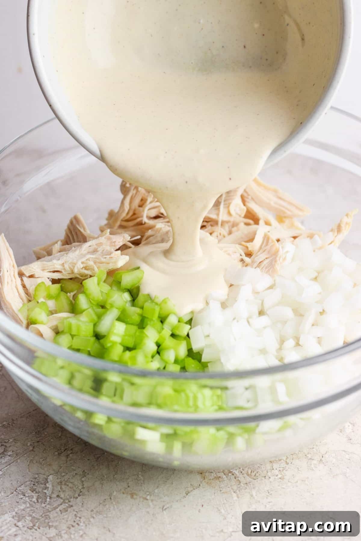 A creamy sauce is being poured into a bowl containing shredded chicken, diced onions, and chopped celery.