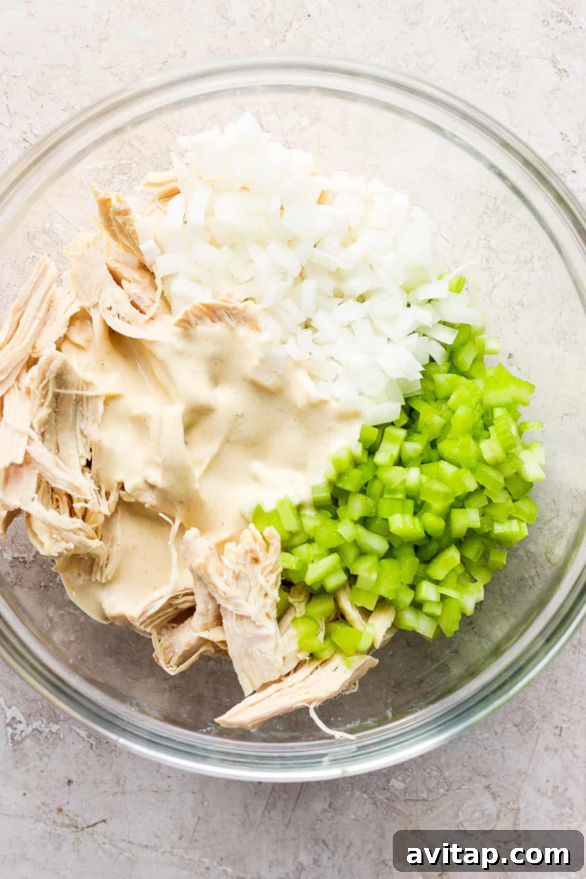 A glass bowl containing shredded chicken, diced onions, and chopped celery, possibly in preparation for a chicken salad or similar dish.