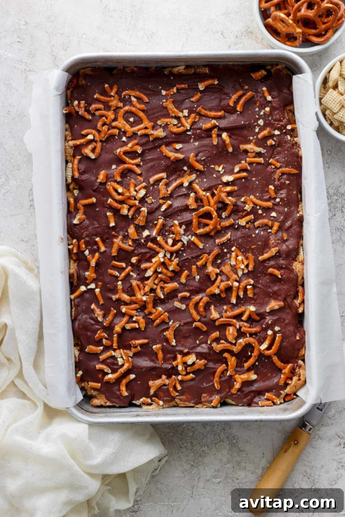 A tray of chocolate-covered toffee sprinkled with pretzel pieces on a kitchen counter, accompanied by a cloth and a spatula.