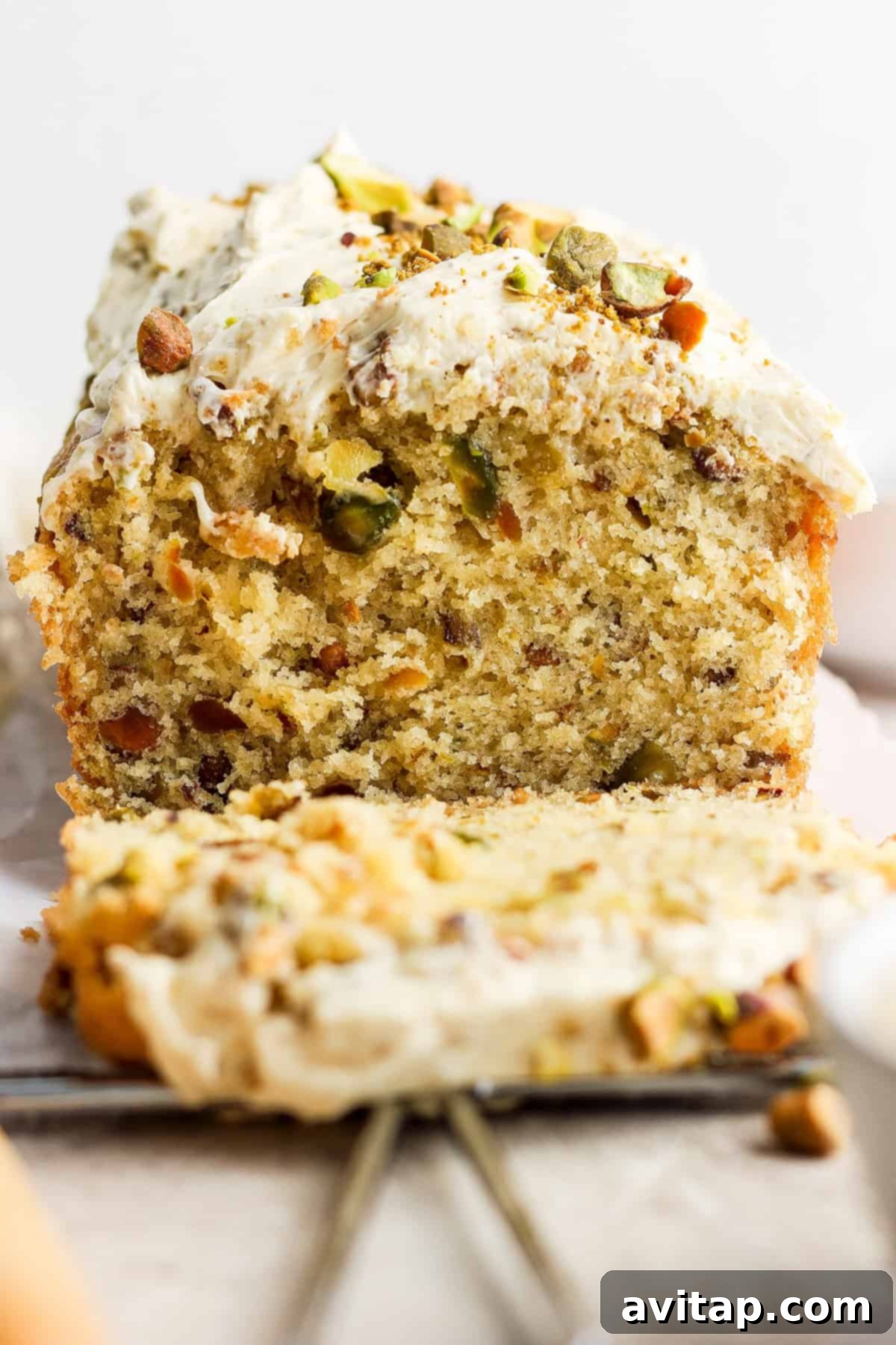 A close-up of a slice of pistachio cake with frosting and chopped nuts on top, with the remaining cake in the background.