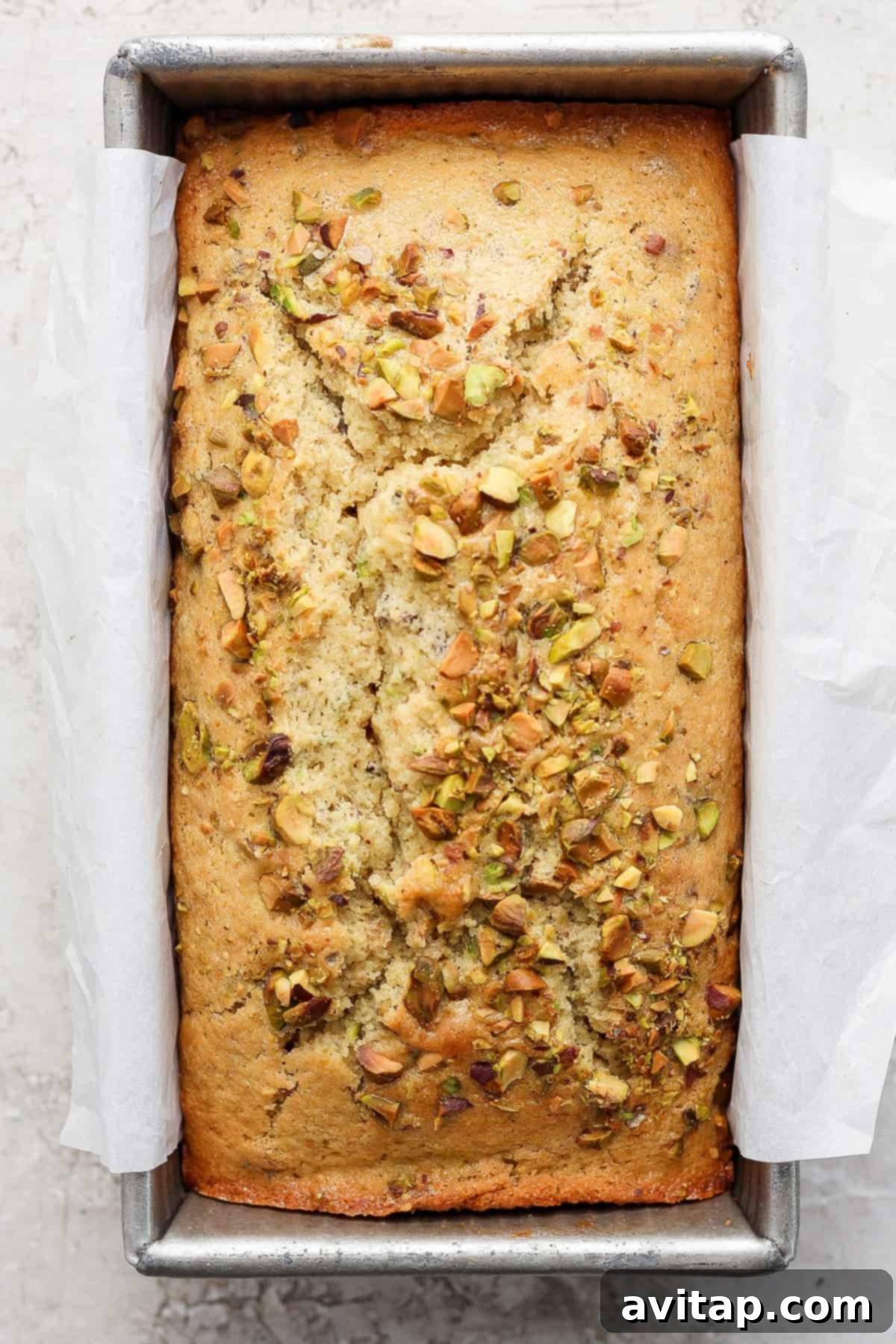 A freshly-baked loaf of bread with pistachios sprinkled on top, sitting in a metal baking pan lined with parchment paper.