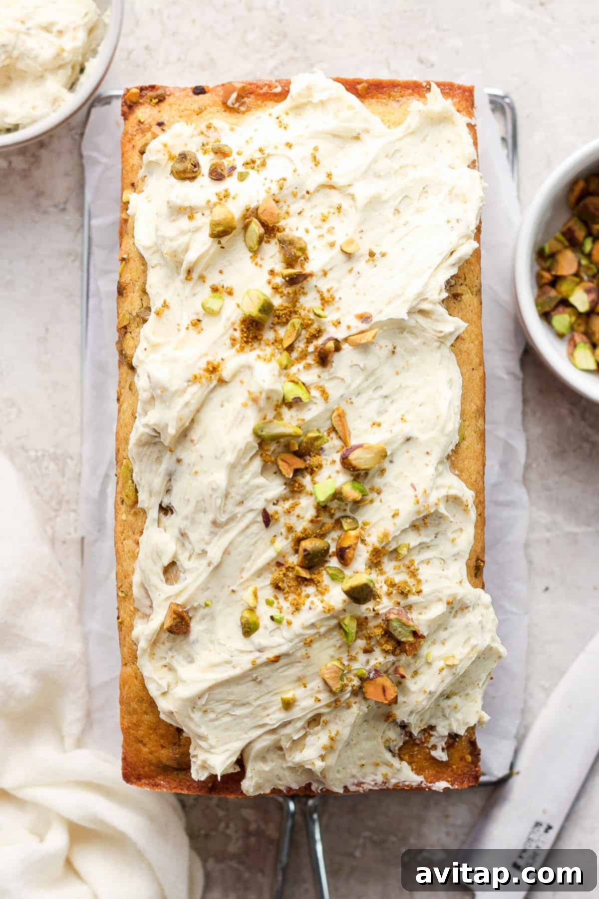 A freshly baked loaf cake topped with a creamy white frosting and sprinkled with chopped pistachios.
