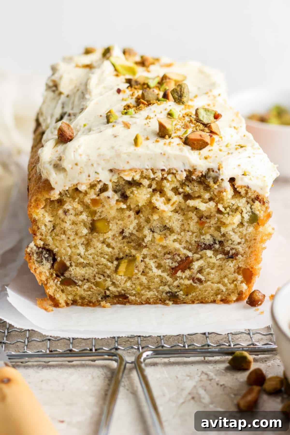 A loaf of frosted cake with nuts and chopped pistachios on top, partially sliced, displayed on a piece of parchment paper.