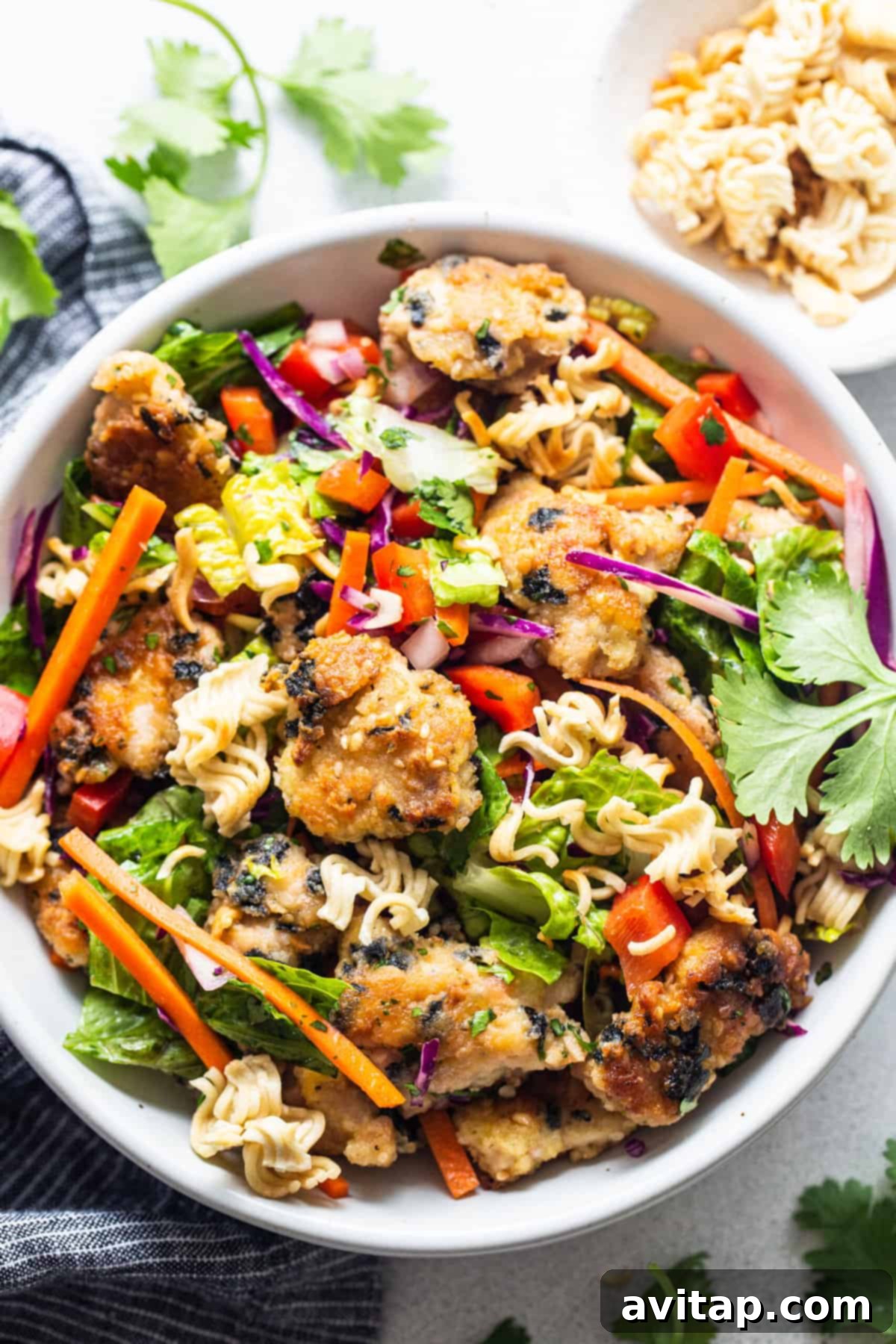 A vibrant ramen noodle salad with crispy nori-coated chicken, fresh vegetables, and toasted ramen noodles in a white bowl, ready to be served.