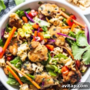 The final presentation of an Asian chicken noodle salad in a white bowl, topped with crispy chicken and ramen, garnished with fresh herbs and sesame seeds.