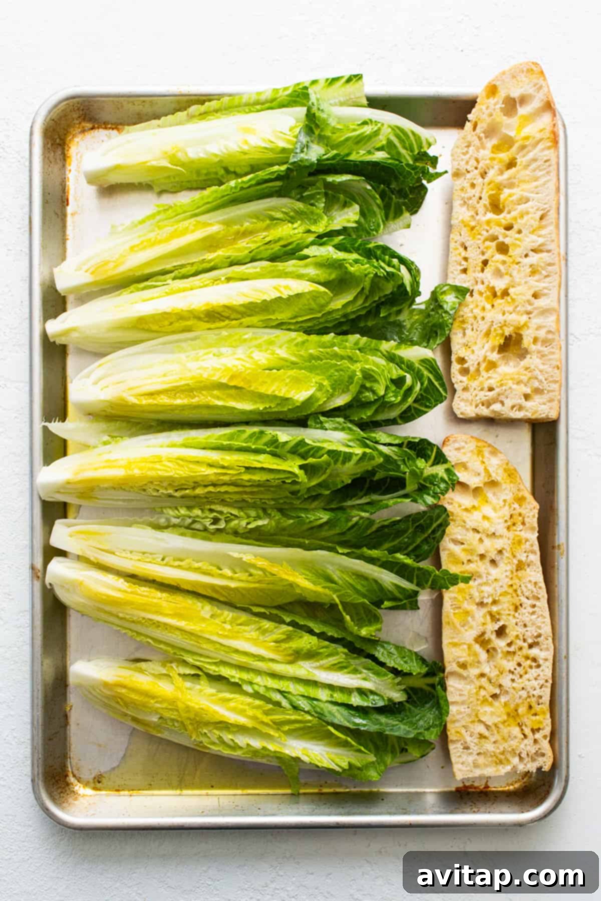 Flame-Kissed Caesar with Crispy Charred Croutons 3 Fresh heads of romaine lettuce and slices of French baguette arranged on a baking tray, ready for grilling.