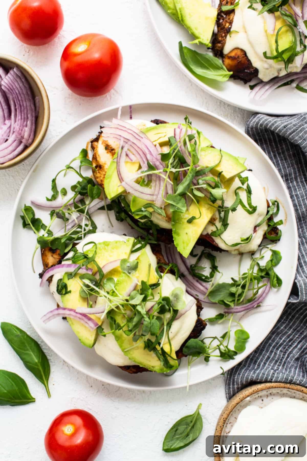 California Sunshine Grilled Chicken 2 Two plates with avocado, tomatoes and onions on them. Each plate features grilled chicken topped with fresh mozzarella, sliced tomatoes, creamy avocado, and red onion, garnished with microgreens and basil.