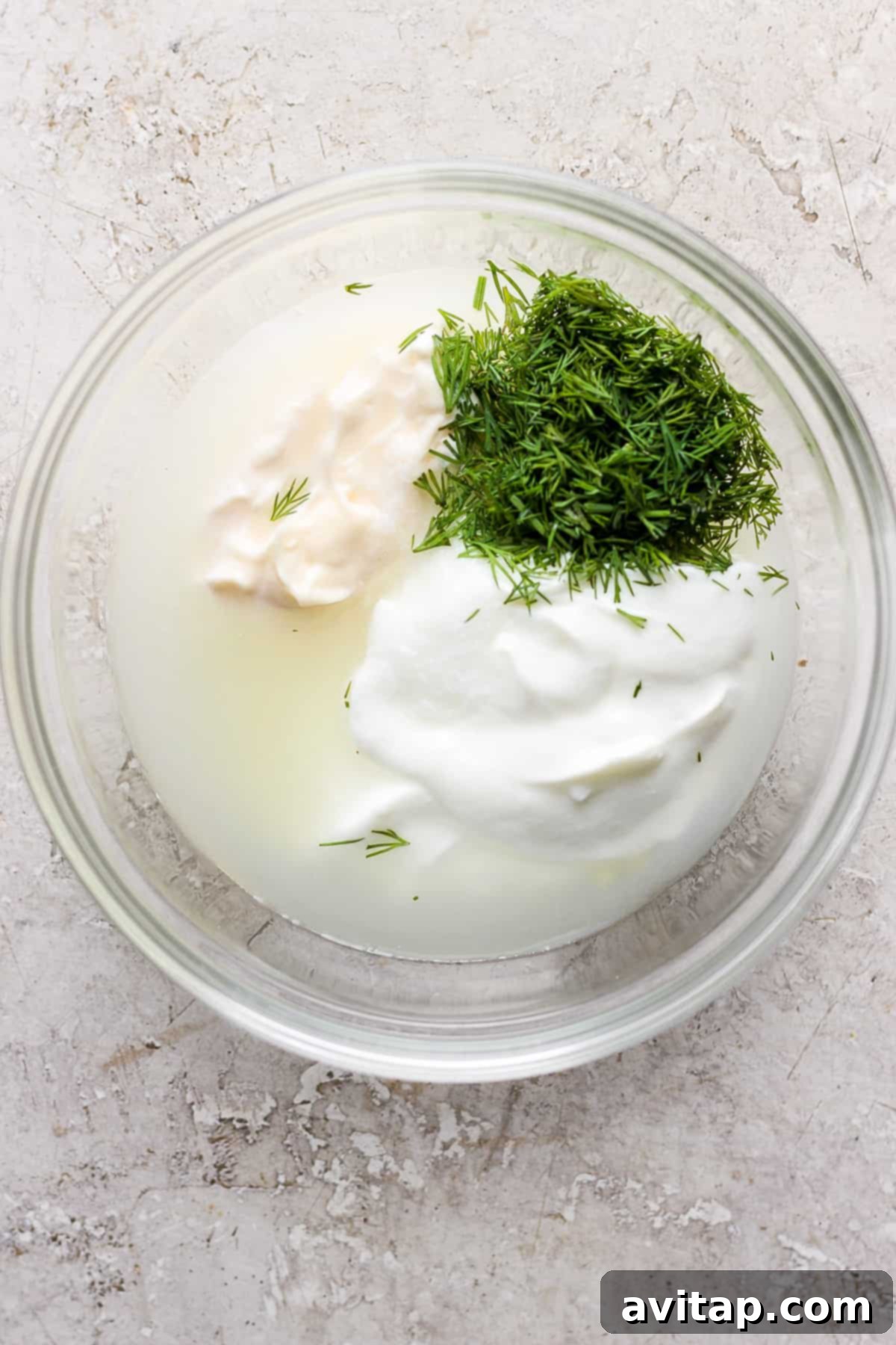 A glass bowl containing sour cream, dill, and a liquid, possibly part of a recipe preparation.