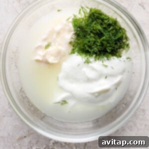 A glass bowl containing sour cream, dill, and a liquid, possibly part of a recipe preparation.
