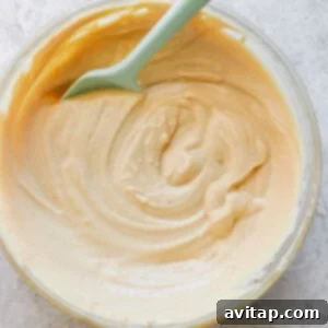 A glass bowl containing creamy peanut butter with a green spoon, photographed from overhead on a textured white surface.