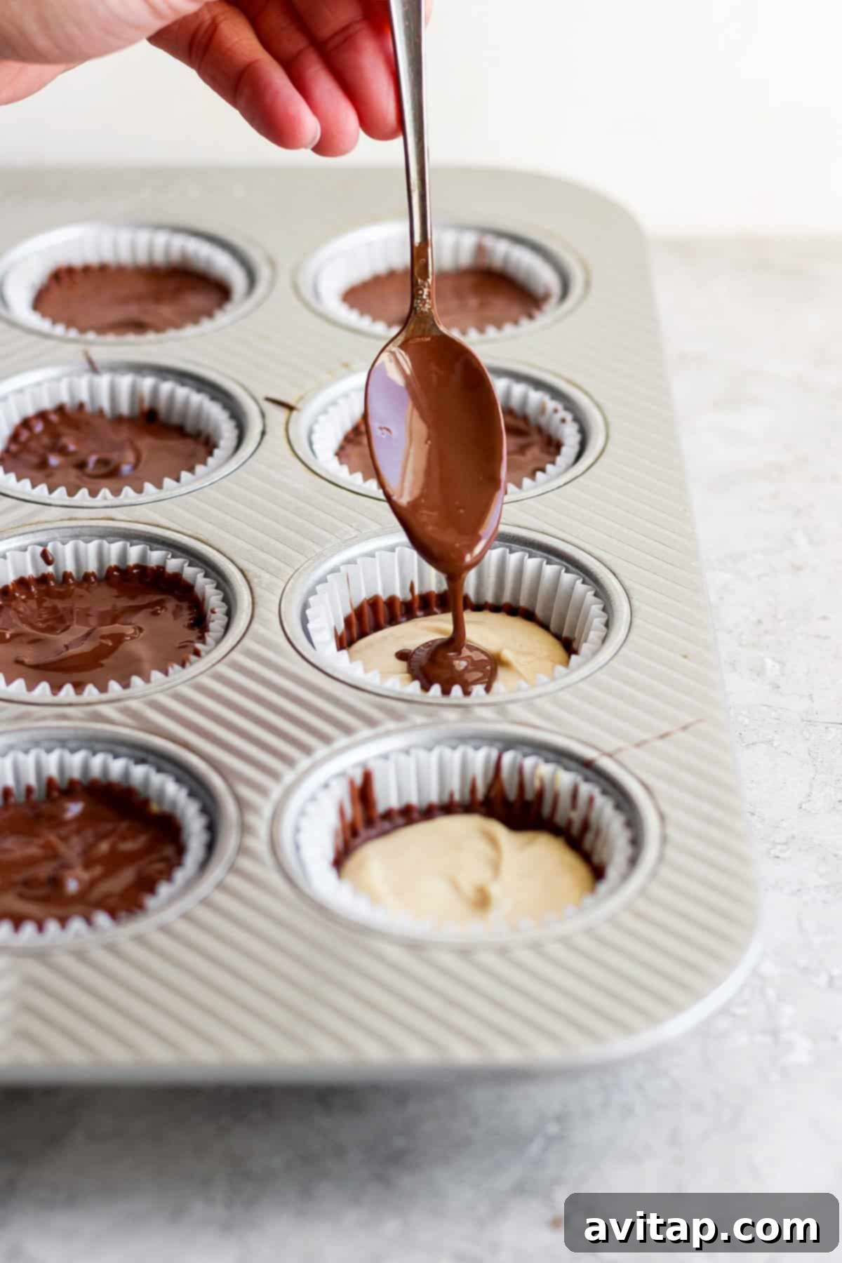 Drizzling chocolate over peanut butter in lined muffin cups using a spoon.