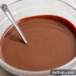 A glass bowl containing smooth melted chocolate with a metal spoon resting inside, set on a light-colored surface.