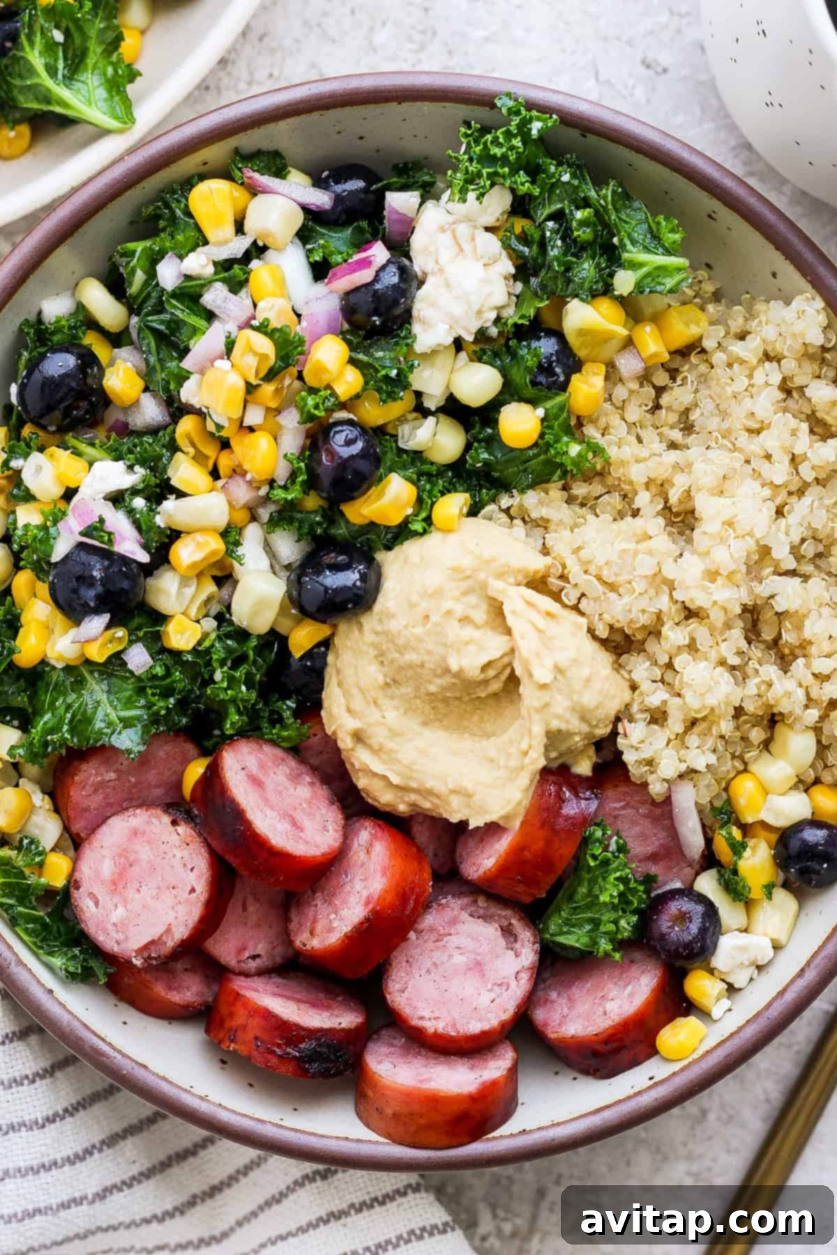 A beautifully arranged protein bowl featuring fluffy quinoa, perfectly sliced sausage, a generous dollop of creamy hummus, a fresh kale salad with blueberries and corn, and other vibrant ingredients, showcasing a wholesome and colorful meal.