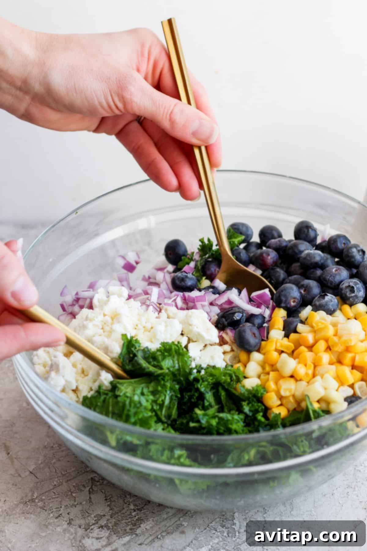 A person uses golden tongs to expertly mix a colorful and fresh salad featuring bright blueberries, crisp corn, chopped red onions, and leafy greens in a clear glass bowl, showcasing the vibrant preparation for the protein bowl.