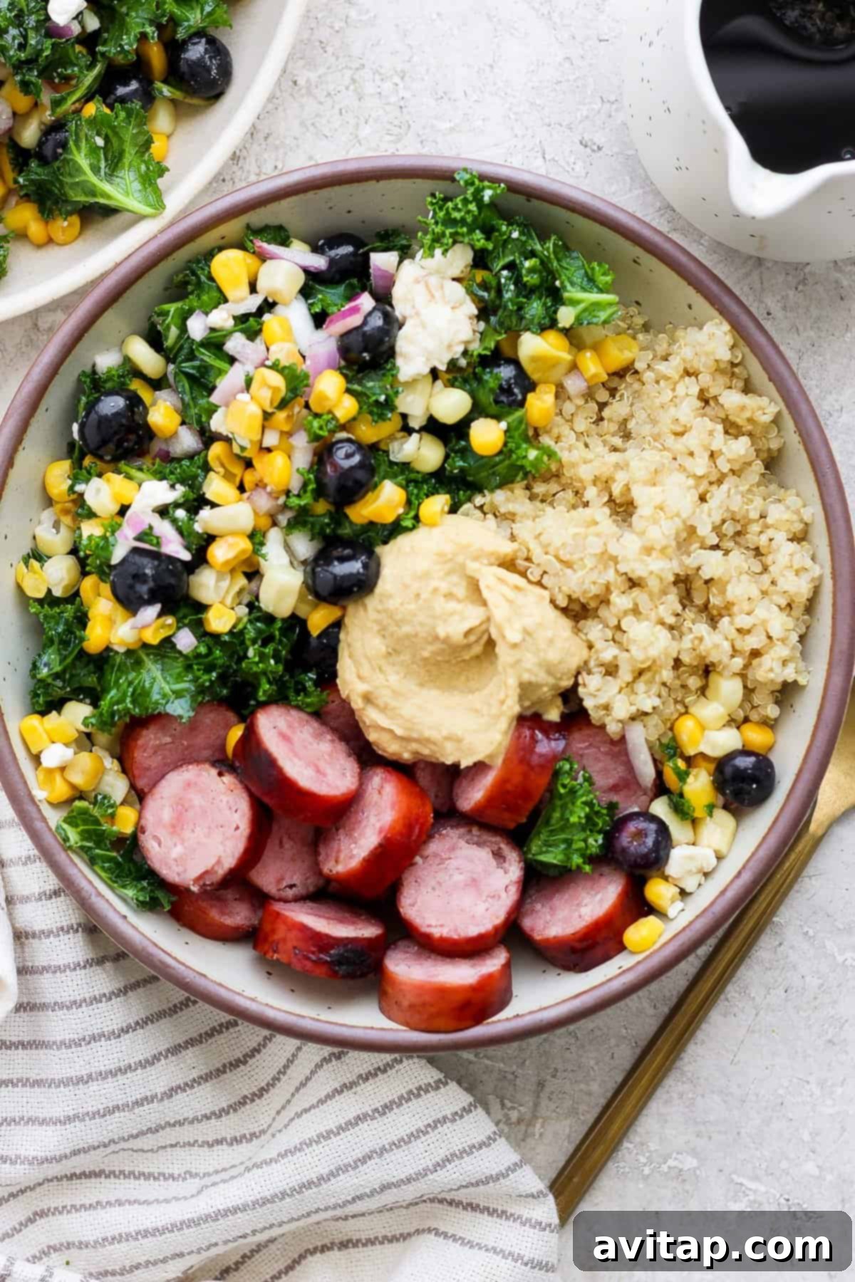 A perfectly portioned protein bowl, showcasing fluffy quinoa, savory sliced sausage, a colorful mix of massaged kale and fresh vegetables, all crowned with a generous dollop of creamy hummus, ready to be enjoyed.